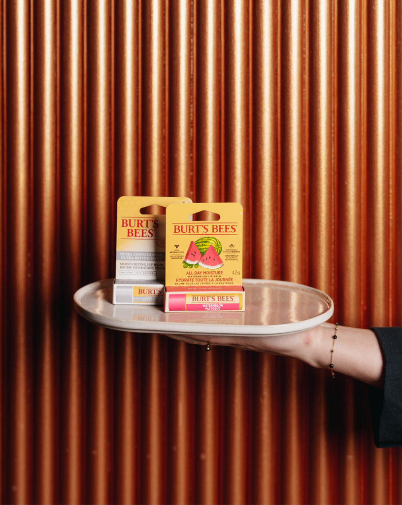 Burt’s Bees lippenbalsems in de varianten Watermelon en Ultra Conditioning, gepresenteerd op een wit dienblad tegen een luxe, koperkleurige achtergrond. Deze 100% natuurlijke lippenbalsems zorgen voor langdurige hydratatie en verzorging. Onderdeel van de &C Voorjaarsbox in samenwerking met Get Your Gift.